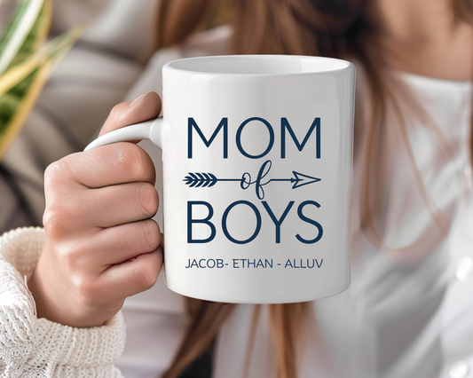White ceramic mug held by a woman in a cozy sweater, with bold navy text reading “Mom of Boys” and personalized names: Jacob, Ethan, Alluv.

